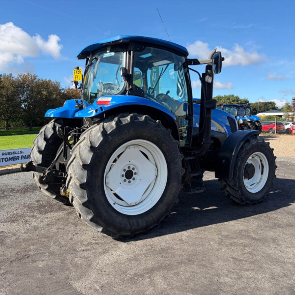 Blue tractor parked outdoors, close-up front view with oversized tires and glass cabin, sign reads 'RUSSELLS CUSTOMER PARKING' nearby