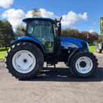 Large blue New Holland tractor on display outdoors, with another tractor in the background under a bright sky.