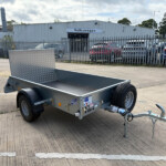 Close-up of a metal utility trailer with a diamond-plate shield, hitched in a parking lot with a corrugated VW building in the background in front of a fence.
