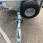 Metal trailer tongue with a mounted spare tire on a gray wheel, attached to a concrete parking lot with a white line nearby.