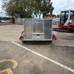 Silver metal trailer with a diamond-plate bed in a paved lot; a small excavator sits to the right with a gate and industrial buildings in the background.