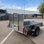 Metal diamond-plate utility trailer in a parking lot near a tall metal fence and industrial building behind it.
