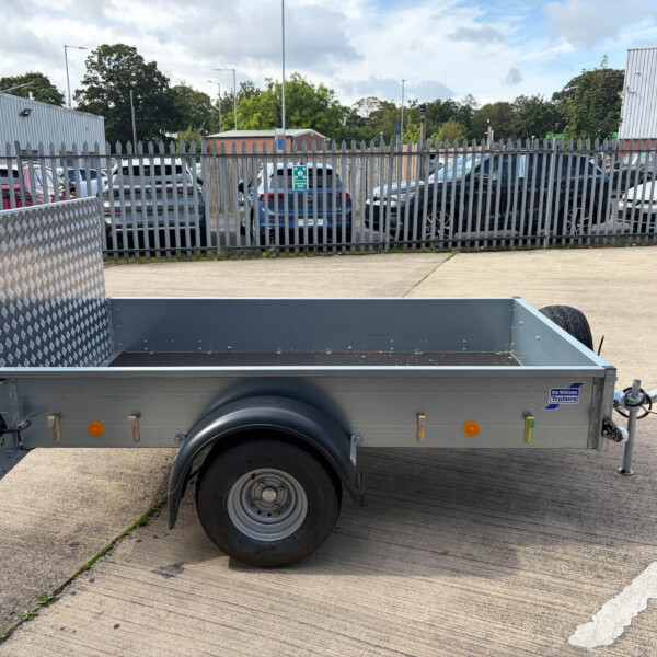 Close-up side view of a gray utility trailer in a parking lot, showing a wheel, hitch, and diamond-plate side panel with a fence and cars in the background