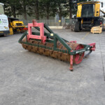 Industrial equipment yard with a red-green soil roller in the foreground and a large yellow excavator in the background, plus a van on the left.