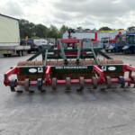 Front view of a heavy red-green cultivator with spikes in a paved yard, NRH ENGINEERING label on the frame, with tractors and industrial buildings in the background