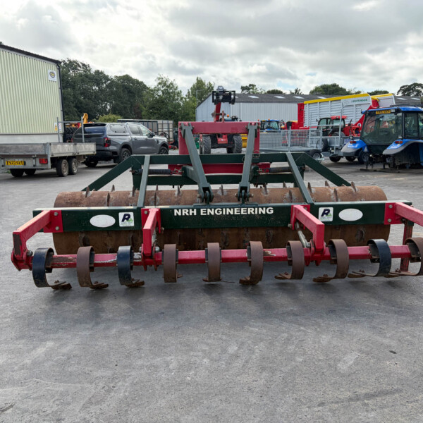 Front view of a heavy red-green cultivator with spikes in a paved yard, NRH ENGINEERING label on the frame, with tractors and industrial buildings in the background
