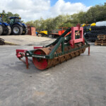 Two blue tractors parked in a farm equipment yard with a large red-green roller in the foreground and pallets nearby.