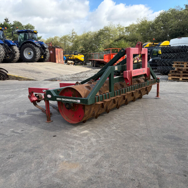 Two blue tractors parked in a farm equipment yard with a large red-green roller in the foreground and pallets nearby.