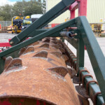 Close-up of a rusted orange agricultural roller attached to a green metal frame in an outdoor yard with pallets and a building in the background.