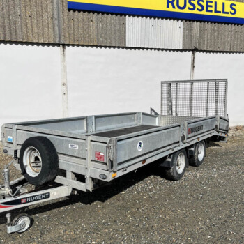 Grey metal double-axle utility trailer on a gravel lot, with a yellow-and-blue Russell sign on a corrugated-wall building in the background.