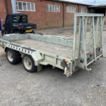 Galvanized flatbed trailer with metal side frames in an industrial yard; a black Polaris Ranger UTV sits on the trailer, brick warehouse backdrop.