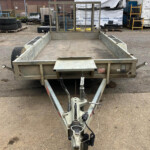 Rear view of an empty metal utility trailer with a mesh gate, parked on a gravel industrial lot.