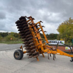 Orange disc harrow parked on a road, discs stacked on a yellow frame under a cloudy sky with trees in the background