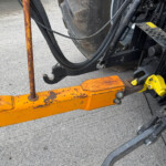 Close-up of a heavy machinery attachment with orange safety bars, black hydraulic hoses, and yellow warning decals on a concrete surface.