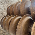 Close-up of rusted, helical auger blades lying in a dirt and straw-covered outdoor area, showing weathered metal surfaces.