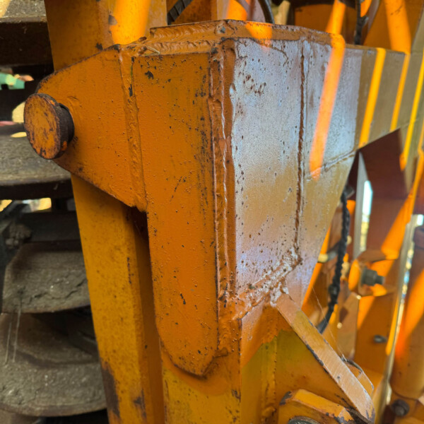 Close-up of orange steel machinery with rust, showing welded joints and a hydraulic pin/bolt assembly on a heavy frame.