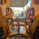 Inner view of large orange industrial machinery with hydraulic cylinders, chains, and metal arms inside a shed. Outside, agricultural equipment and a car are visible under a corrugated roof.