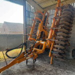 Orange disc harrow machinery stacked inside a shed, with large round metal discs and hydraulic lines visible.