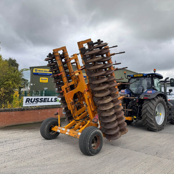 Orange disk harrow attached to a blue tractor parked in front of an agricultural equipment shop with Russell’s sign visible under cloudy skies.