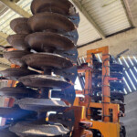 Rusty circular harrow discs mounted on an orange frame inside a shed, an agricultural implement awaiting use.