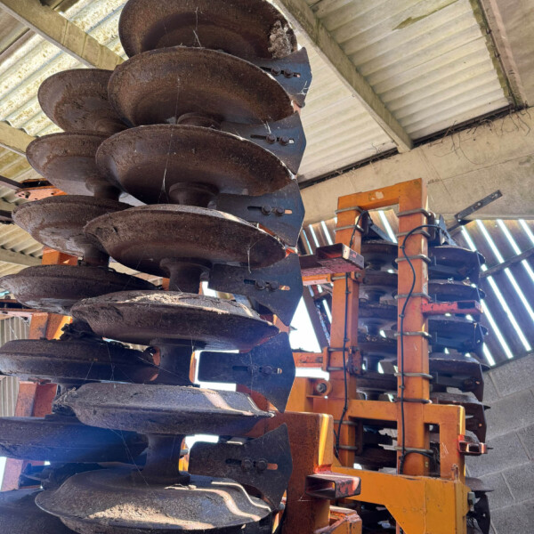 Rusty circular harrow discs mounted on an orange frame inside a shed, an agricultural implement awaiting use.
