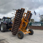 Blue tractor with an orange multi-disc harrow attached, on a dealership lot with flags in the background