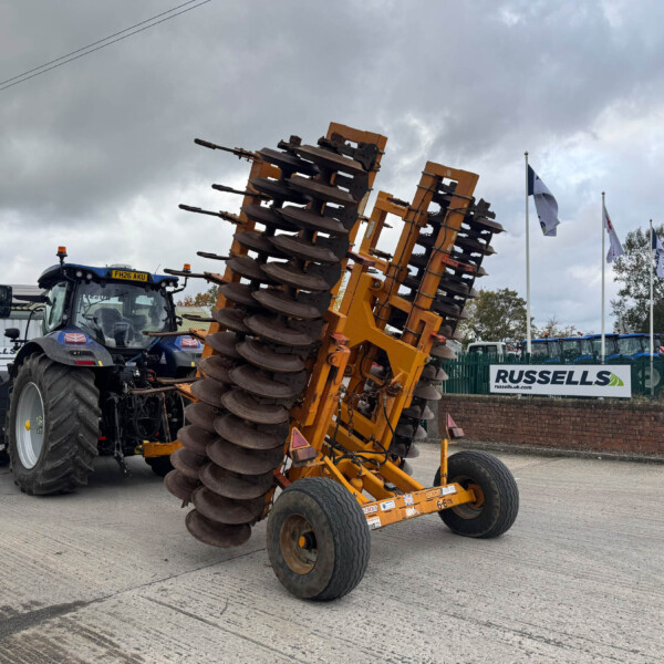 Blue tractor with an orange multi-disc harrow attached, on a dealership lot with flags in the background