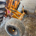 Orange agricultural implement with large, worn tires and rusted metal components leaning against a concrete block wall.