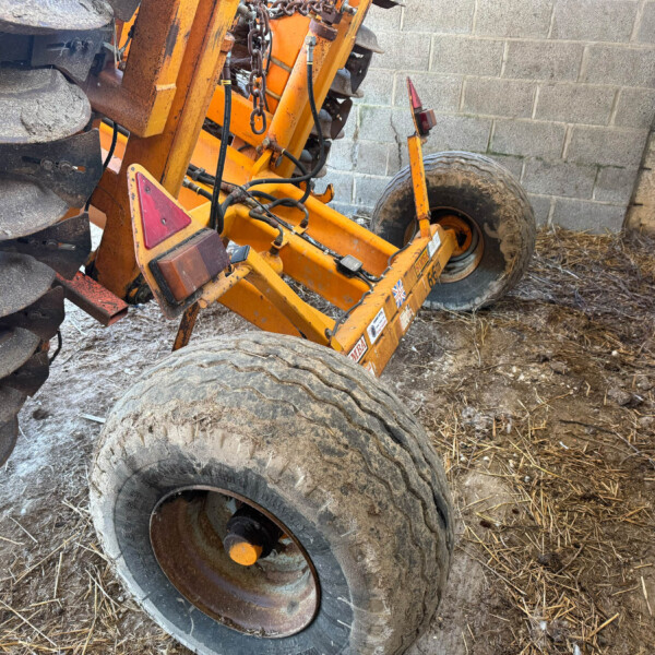 Orange agricultural implement with large, worn tires and rusted metal components leaning against a concrete block wall.