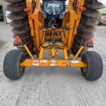 Rear view of an orange farming implement with large circular discs on both sides, hydraulic lines, and warning triangles in a yard.