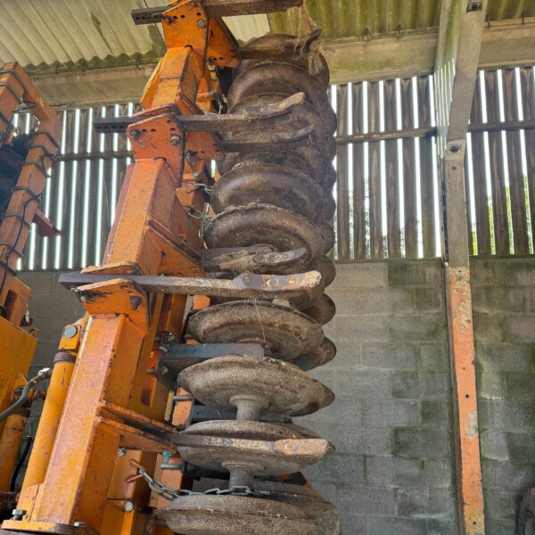 Rusty orange disc harrow attachment with multiple round discs mounted on a frame inside a shed.