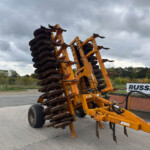 Large orange multi-disc farming implement tilted on a street under a cloudy sky, with a Russell sign visible in the background.