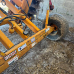 Orange farming implement frame with hydraulic hoses and a large dusty tire, parked beside a concrete block wall; labels read SIMBA and warning stickers visible on the frame.
