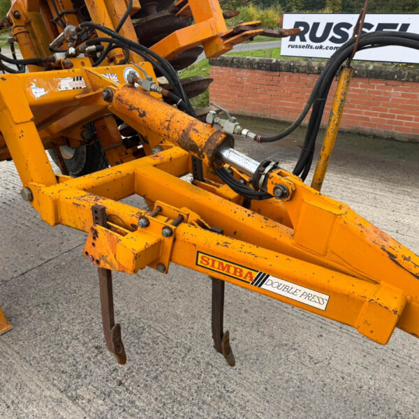Close-up of orange hydraulic construction frame with hoses and metal joints, showing a 'DOUBLE PRESS' label and a brick wall in the background.