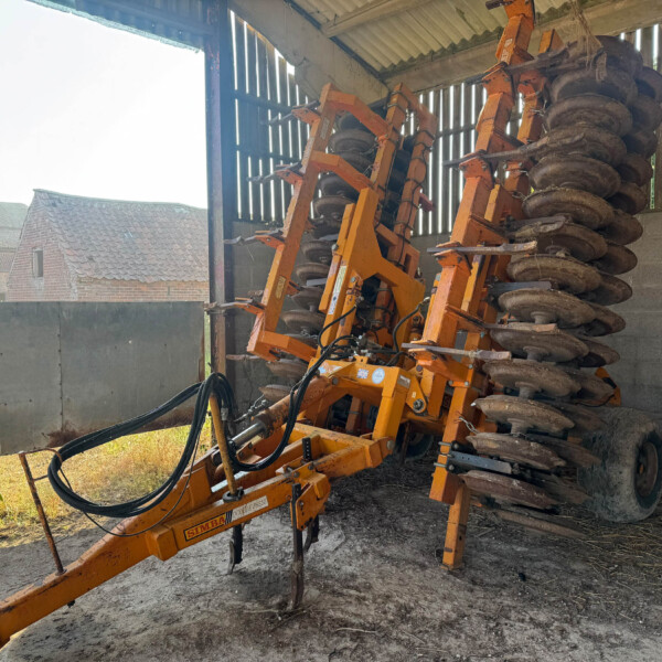 Orange disc harrow machinery stacked inside a shed, with large round metal discs and hydraulic lines visible.