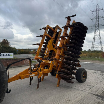 Yellow disc harrow farming implement with multiple rotating discs, mounted on a frame near a road on a cloudy day