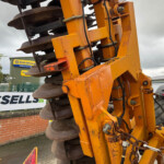 Close-up of an orange road roller/soil compactor with large metal drums and hydraulic components at a construction site, sign visible in background.