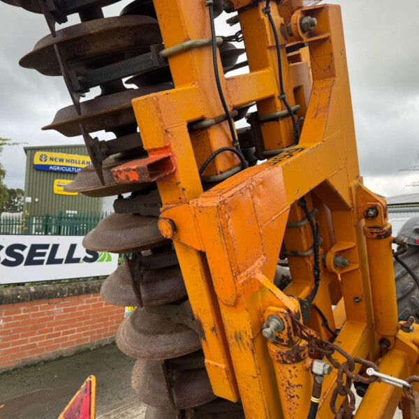 Close-up of an orange road roller/soil compactor with large metal drums and hydraulic components at a construction site, sign visible in background.