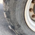Close-up of a dusty tire and rusty white wheel, showing worn tread and rusted lug bolts on the rim.