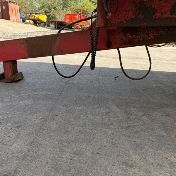 Rusted red steel beam and hitch resting on a concrete ground with hoses hanging from it, in an outdoor yard with machinery in background