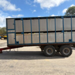Blue metal-framed cargo trailer on wheels parked on a concrete lot with a bright blue sky and clouds above.