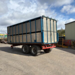Blue industrial trailer on a wheeled platform in a construction yard with equipment in the background, under a cloudy sky.