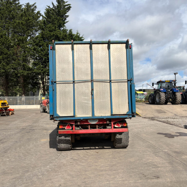 Rear view of a blue trailer in an industrial yard, with tractors lined up to the right and a white van to the left behind a fence.