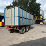 Large blue trailer loaded with tall metal panels in an industrial yard, with a yellow excavator and silos in the background, under a cloudy sky.