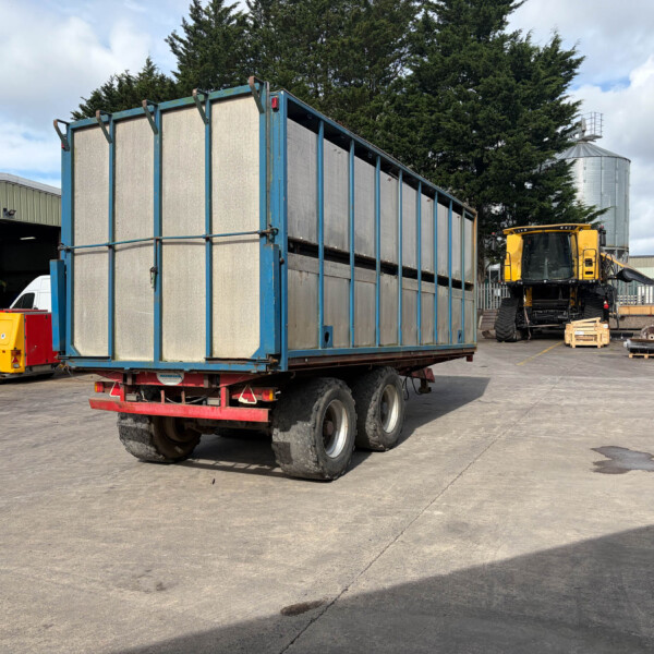 Large blue trailer loaded with tall metal panels in an industrial yard, with a yellow excavator and silos in the background, under a cloudy sky.