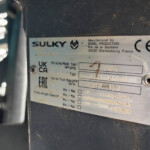 Close-up of a metal machinery label showing the Sulky brand, a circular logo, and a CE mark on a dark surface, with small printed manufacture details nearby