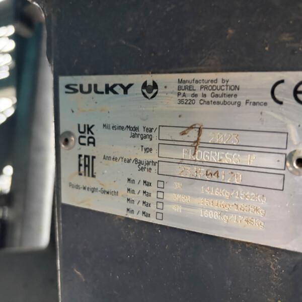 Close-up of a metal machinery label showing the Sulky brand, a circular logo, and a CE mark on a dark surface, with small printed manufacture details nearby