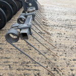 Close-up of a farm or construction implement with curved tines and a gray metal frame on a dirt surface at a work site