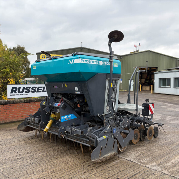 Blue Progress P100 agricultural seeding machine with spiked rollers, parked in an outdoor lot beside a green warehouse.