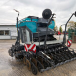 Large blue-green disc cultivator on a trailer in a yard beside a grey metal building.